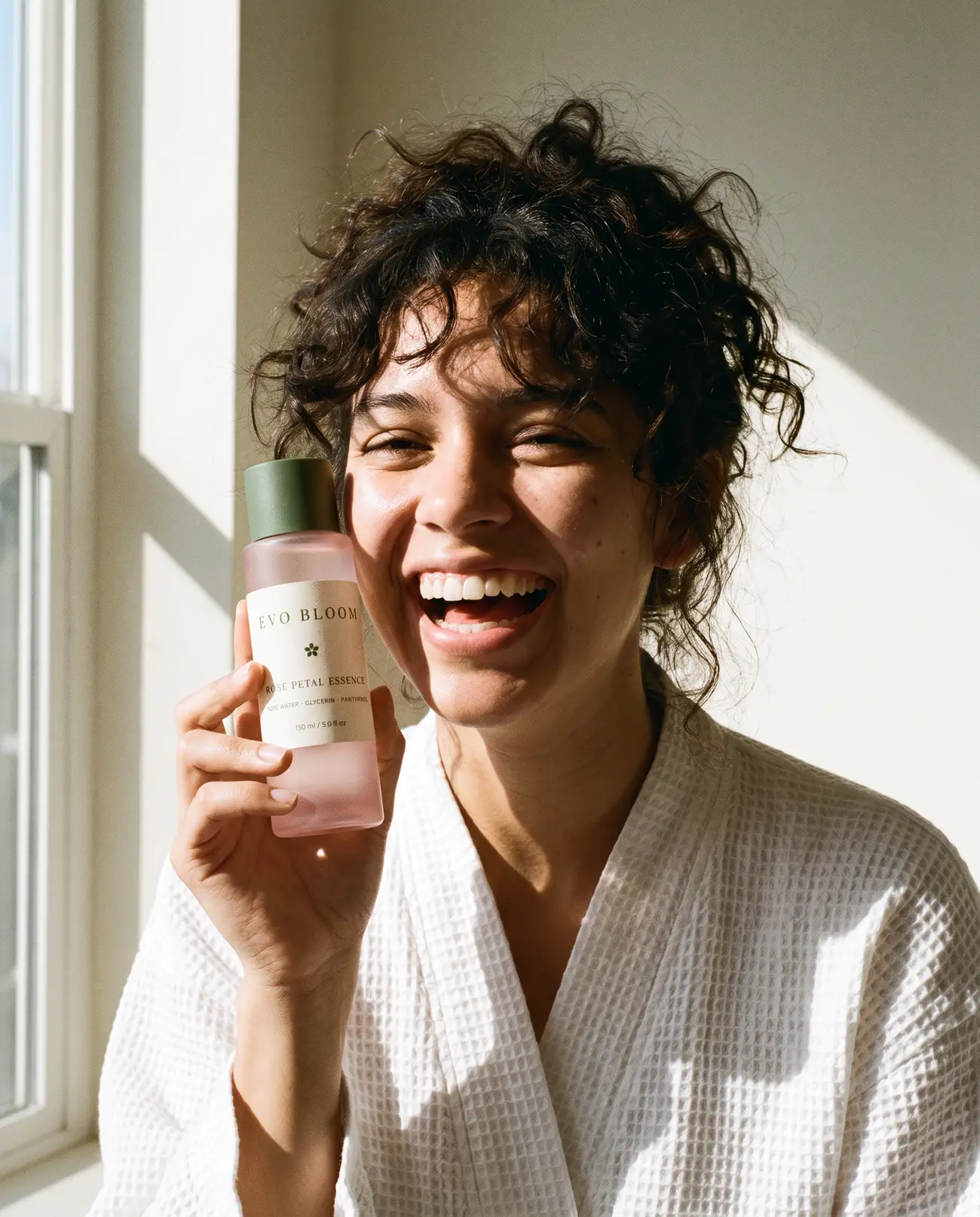 A woman in a white robe laughing, holding a bottle of Rose Petal Essence beside her cheek